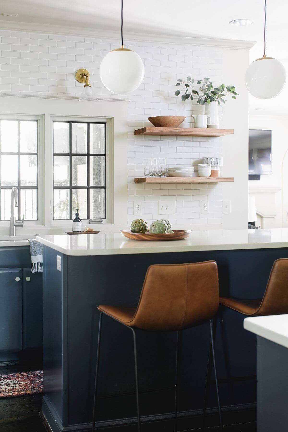 This gleaming white kitchen is accented by rich blue cabinetry below, leather bar stools, handsome lighting and wooden floating shelves. Image: Graham Yelton